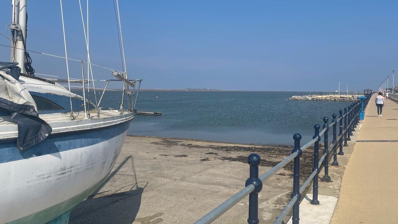 A blue and white boat stands on the beach near the sea on a sunny day at West Kirby, as a person walks past on a walkway, which is bordered by blue and grey iron railings