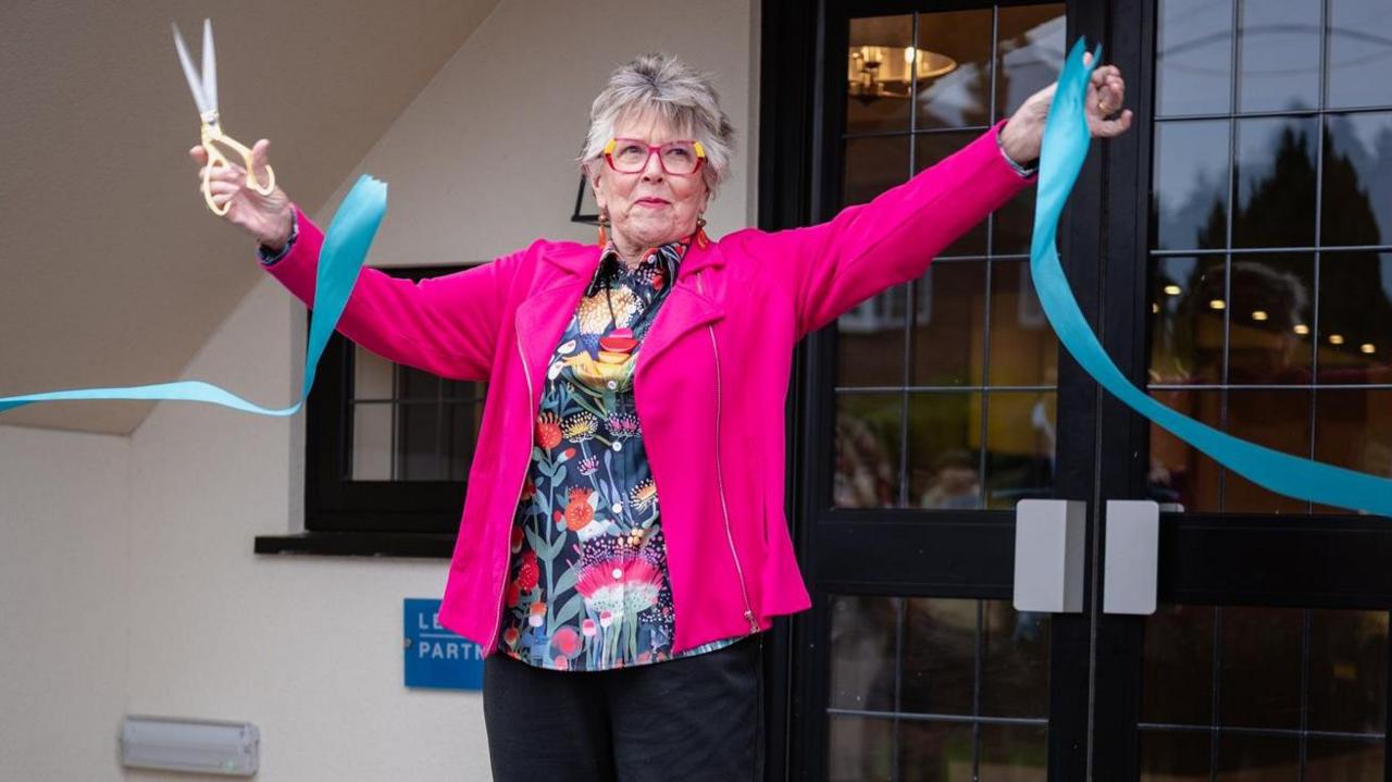 Prue holds her arms aloft as she cuts a turquoise ribbon. She holds scissors in her right hand and the ribbon in the left hand. She stands in front of double doors which are black with glass in the middle.