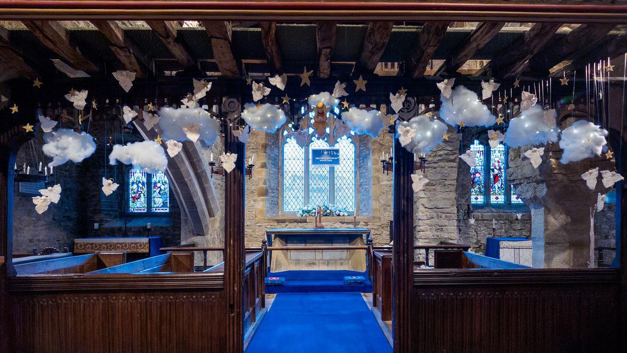Interior of a historic stone church adorned with decorations. Above the altar, cotton-like clouds, knitted angels, and golden stars hang from wooden beams.
