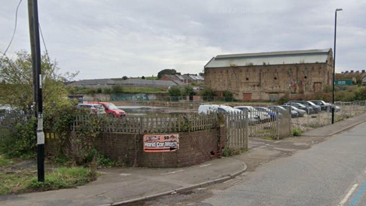 A derelict area of land off Philadelphia Lane. A wall and metal fencing surrounds the area which is being used as a car park. Behind it stands a large, barn-like brick building with pointed roof.