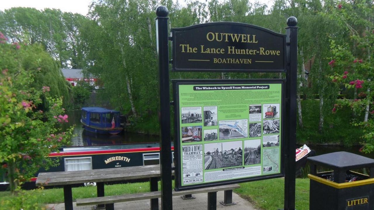A black sign is on the right of the image in front of a small river. It says on it OUTWELL The Lance Hunter-Rowe BOATHAVEN. A picnic bench is behind the sign and a black bin is on the right of it.