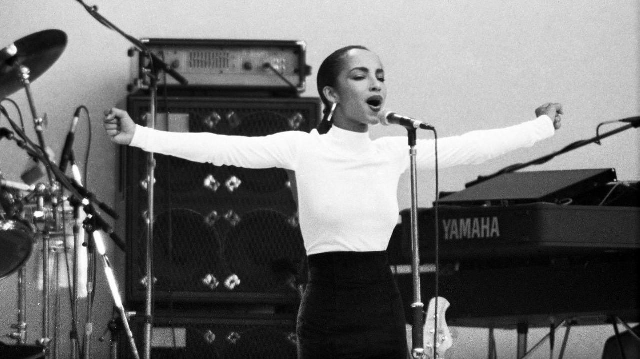 Sade Adu wearing a white polo neck top, standing with her arms outstretched at the microphone. There's a Yamaha keyboard and an amp behind her.
