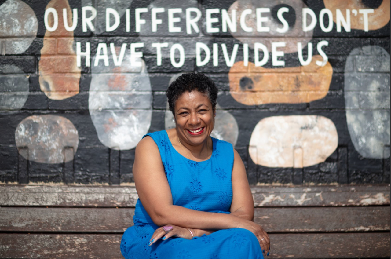 Patrice is seen sitting on some steps with the text 'Our differences don't have to divide us' seen painted on a wall behind her. She is wearing the same blue dress as the previous image.