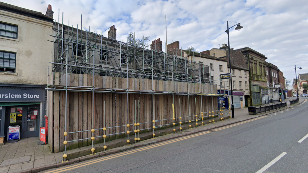 A building in a bank of businesses on a main road which has been completely covered with scaffolding and wooden boards.