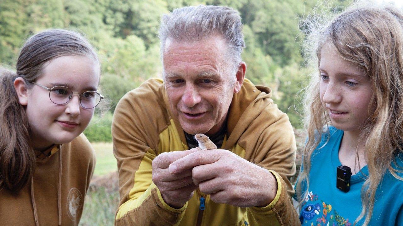 Emily, Eva and Chris Packham look at a tiny harvest mouse sitting on Chris's hand.