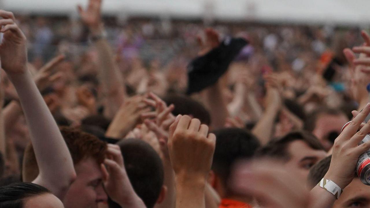 A generic photo of a crowd at an outdoor festival. People's hands are raised in the air.