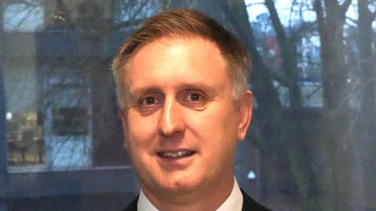 Ed Garrett smiles at the camera as he stands in front of a window. He has short light hair with some grey in it. He wears a black suit with a white shirt. 