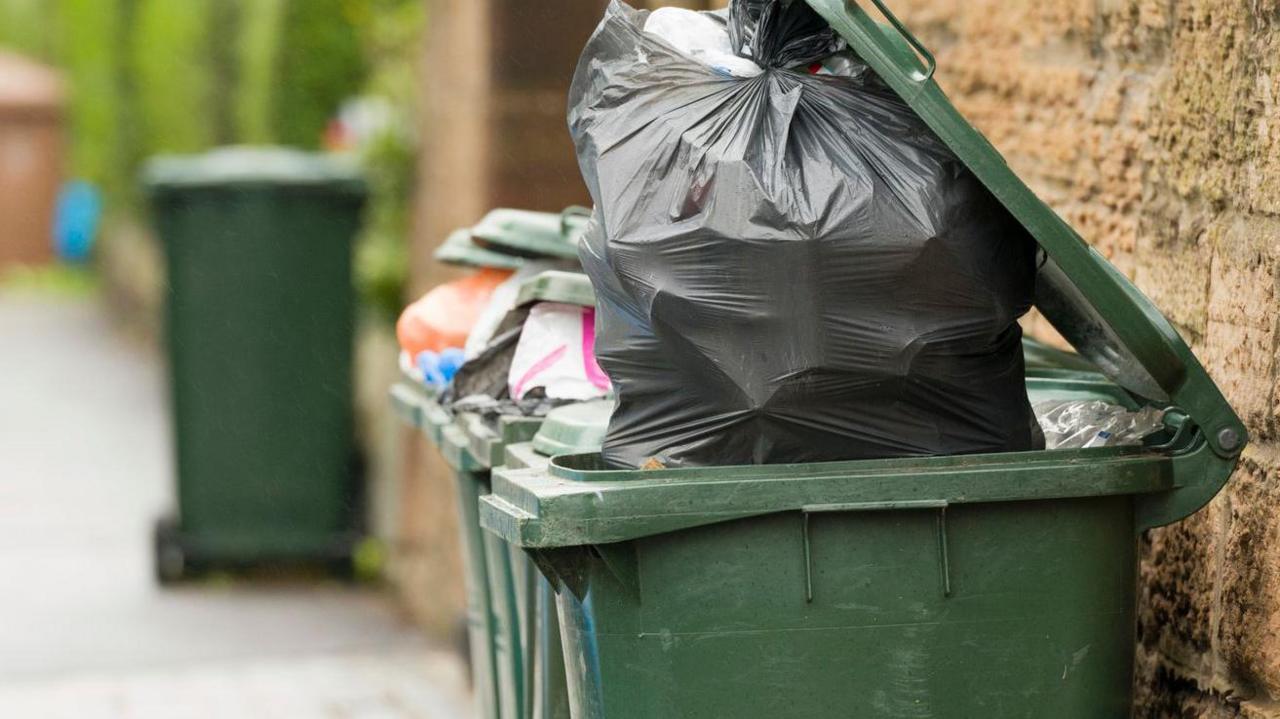 A green wheeled bin full of rubbish with a black bin bag propping open the lid