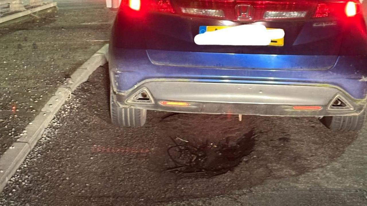 A blue car is stopped at some traffic lights at a roundabout at night. There is a pothole next to its back left tyre and stones on the curb at the side of the road. Inside the pothole looks to be some black wires. 