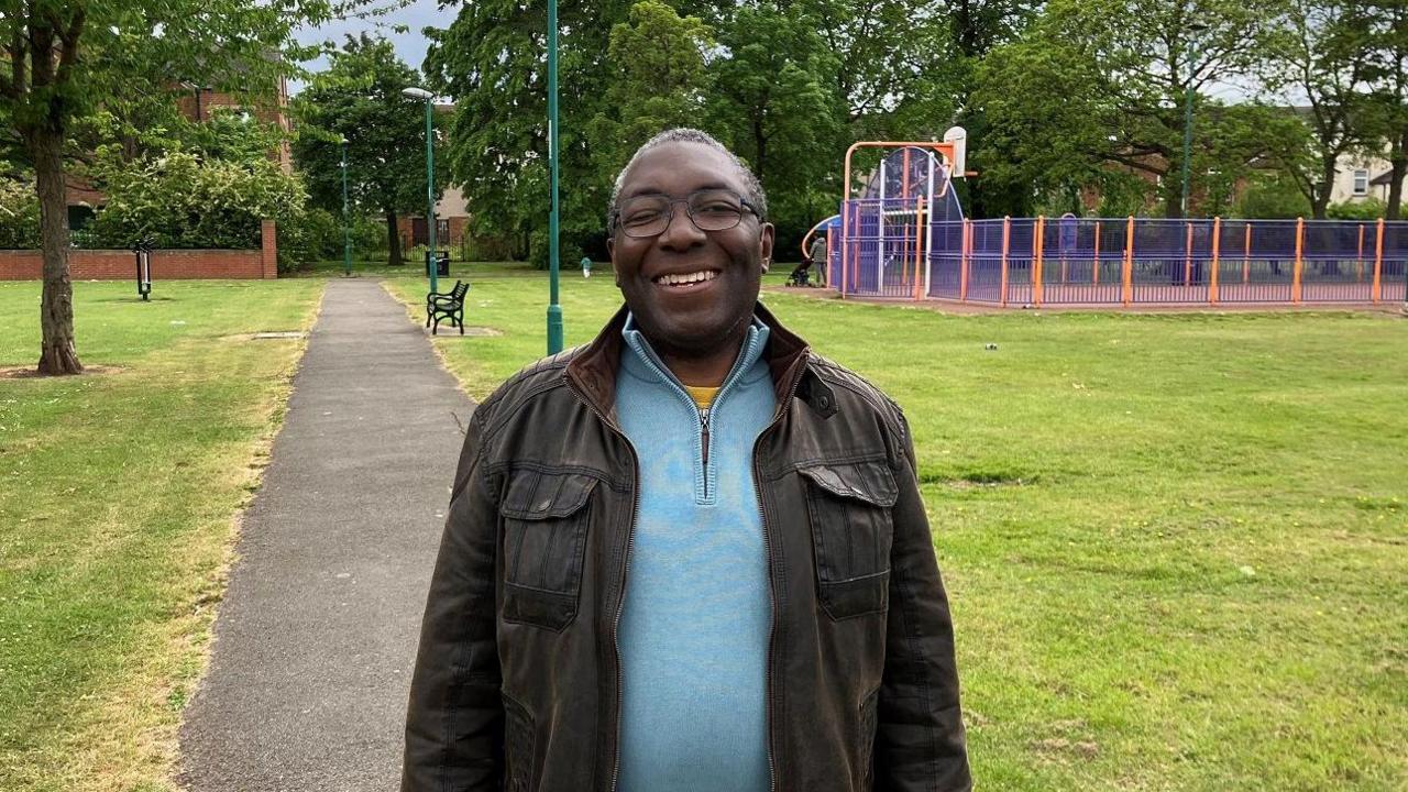 Dominic Nelson-Ashley standing in Ayresome Gardens. He is smiling as he looks into the camera. He has short greying hair and is wearing black glasses, a brown jacket and blue top. Behind him are grassed areas and a basketball court.
