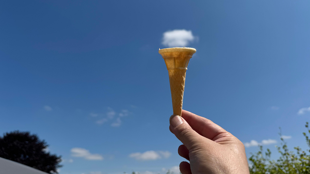 A person is holding an ice cream cone up to an almost totally clear blue sky. A single, small, white cloud is just above the cone, resembling a scoop of ice cream