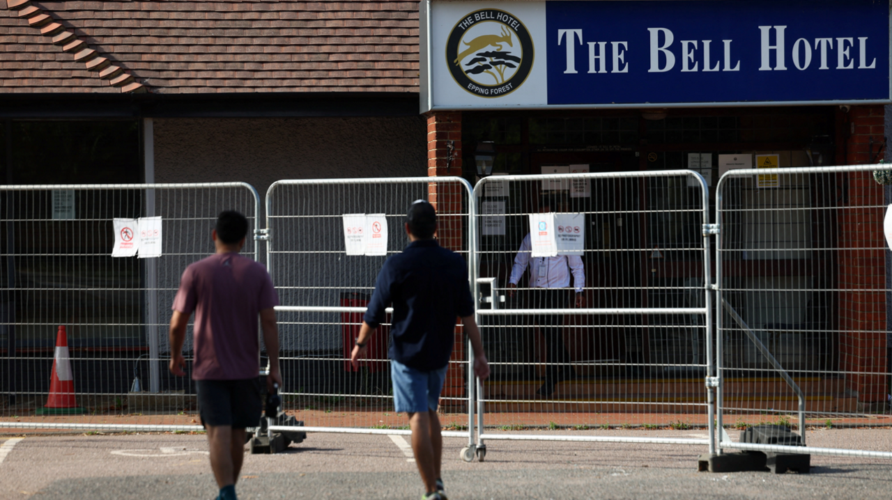 Two men wearing T-shirts and shorts, one has a rucksack they are walking towards a hotel building covered by a metal barrier. Both have their backs to the camera.