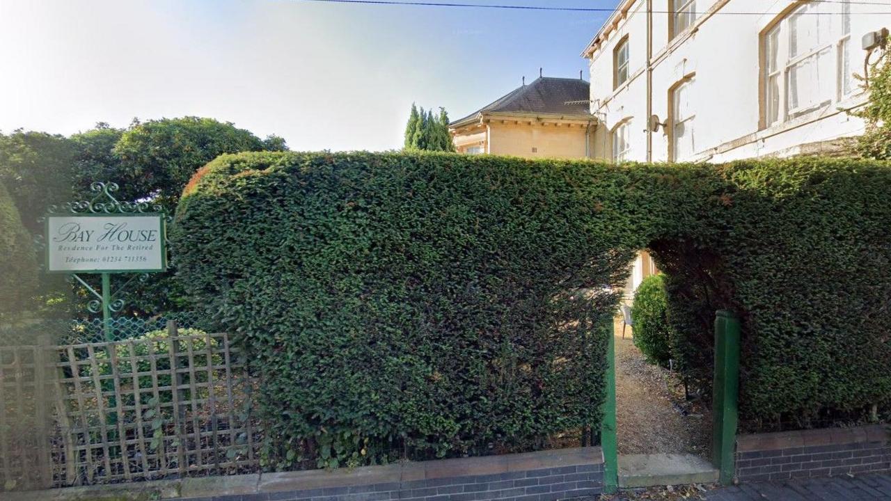 An image of a large hedge in front of a large white building. There is a sign "Bay House" above a fence. 