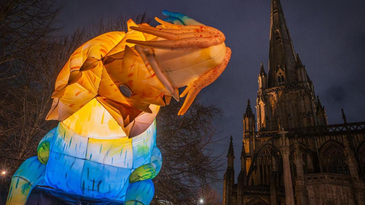 A creature similar to a dragon is rising up near a church. It is lit up from within in different colours, orange, yellow and blue. It is night time behind it. 