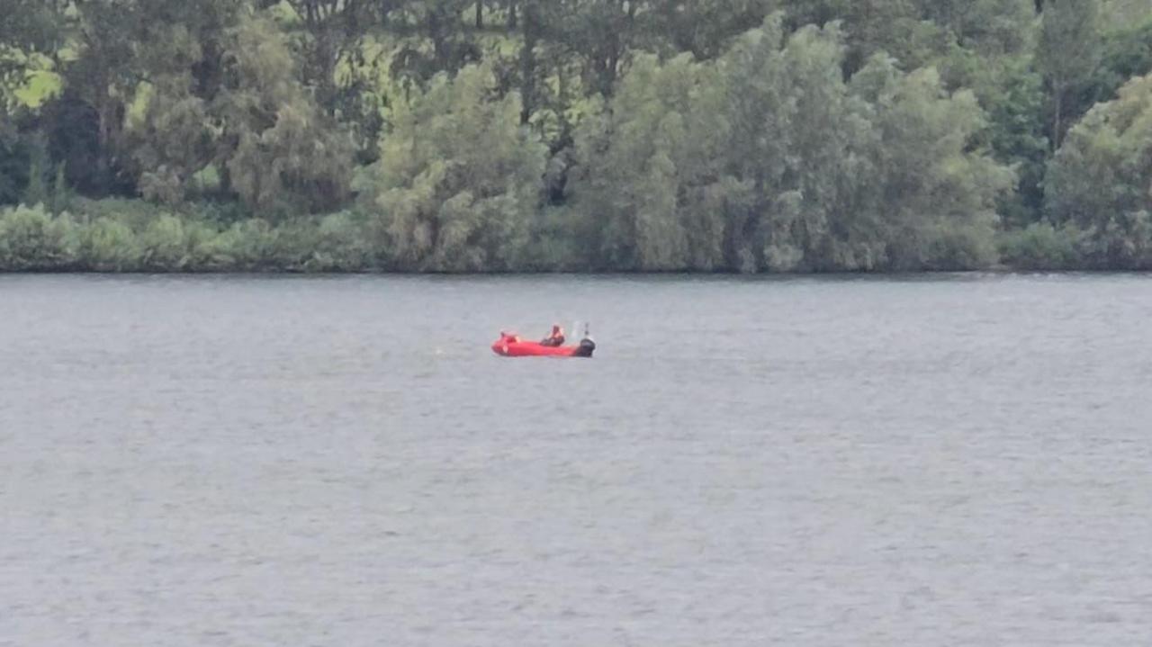 A red rescue boat with a person in it on a reservoir. Trees are seen behind the water.