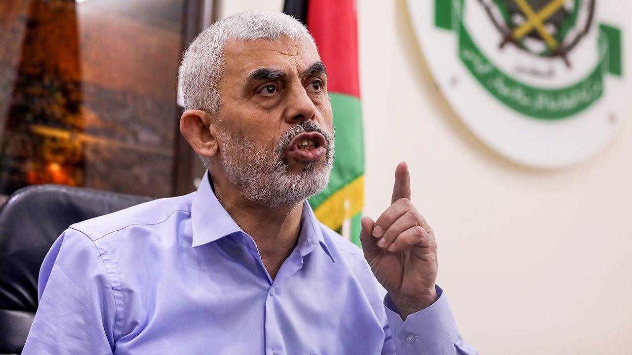 Yahya Sinwar gestures as he speaks while looking away from camera, wearing a purple shirt with grey stubble and hair, with the Palestinian flag and Hamas logo in the background, during a press conference in Gaza City in May 2021