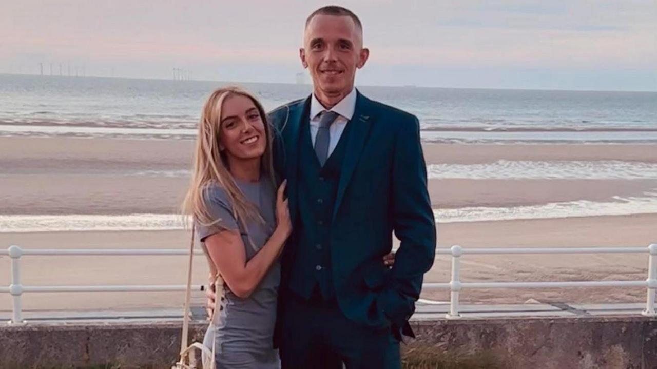 Ryan, dressed in a three piece navy suit and tie, has short brown hair and smiles with his partner, who has blonde long hair and wears a grey dress and cream crossbody bag on her shoulder. Both stand in front of a beach. 