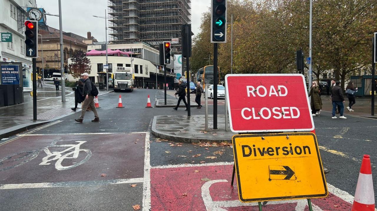 The A38 St Augustine Parade is shut northbound after a burst water main. The road has a road closed sign and a diversion sign with orange cones showing you cannot drive down it. Pedestrians can be seen walking across the closed road in the background