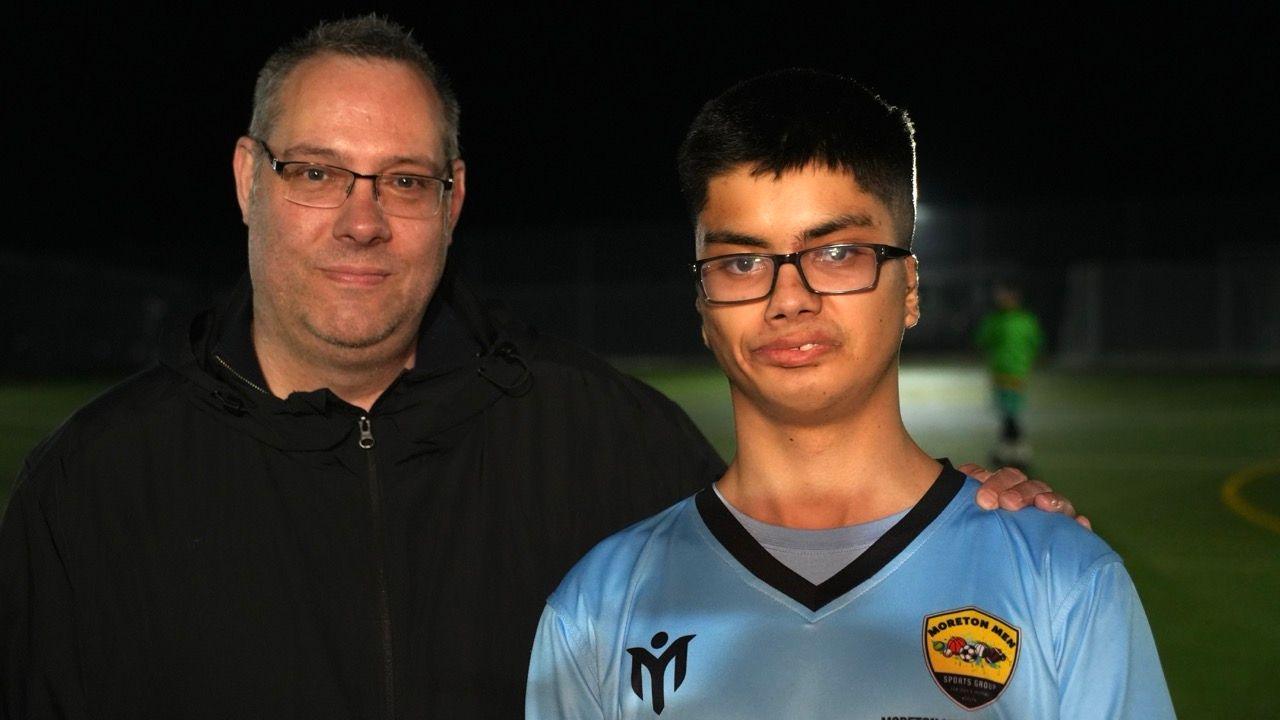 Alex is wearing glasses and the blue Moreton Men football shirt. He has black hair. His dad Damian has his arm round his shoulder. He is wearing a black coat, has greying hair and glasses.
