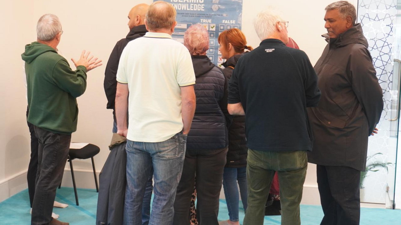 There are several people standing in Masjid Maryam in Acklam. The mosque has bright white lights on the ceiling and a light blue floor. The people look to be in discussion with each other during the mosque's opening day.