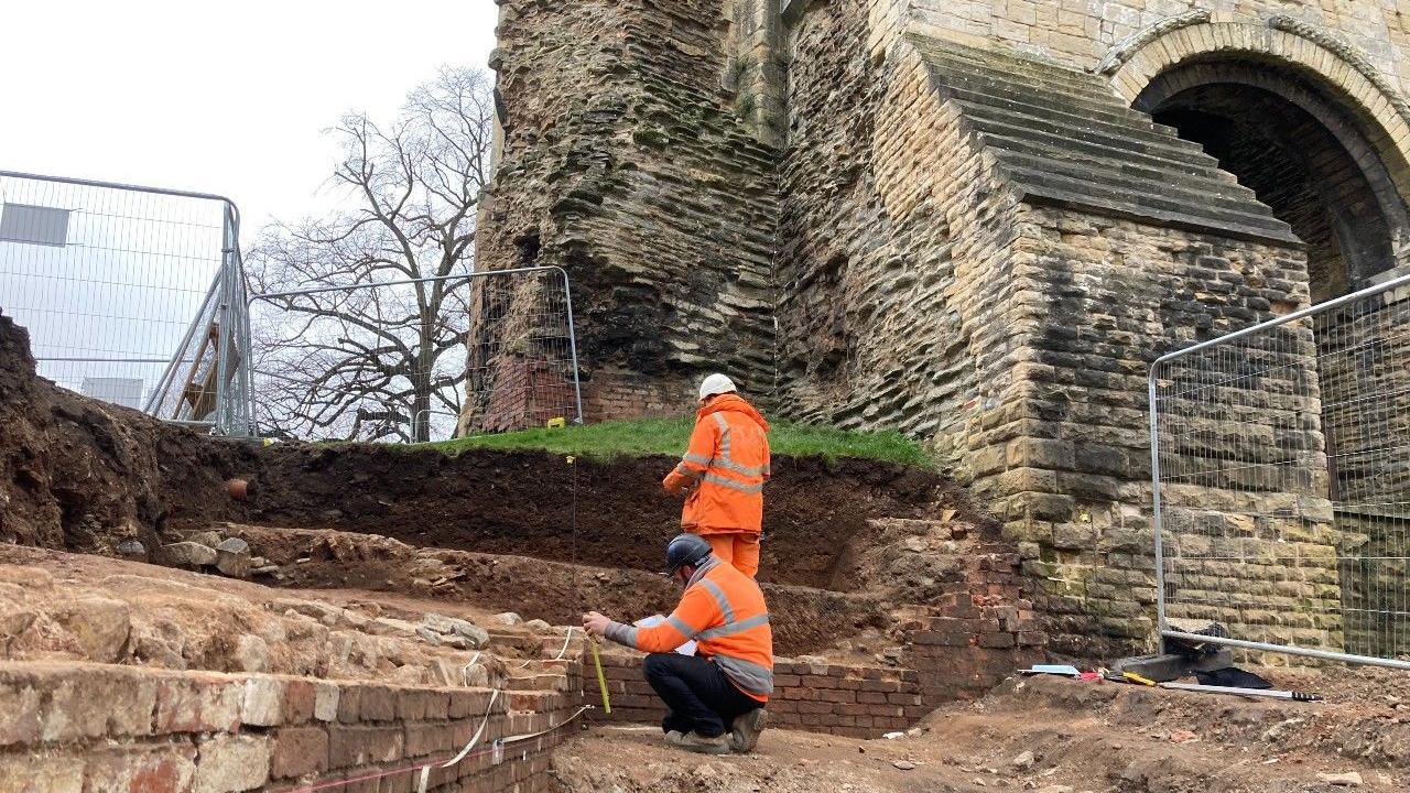 Two archaeologists working outside the castle gatehouse, measuring an exposed stretch of brick wall