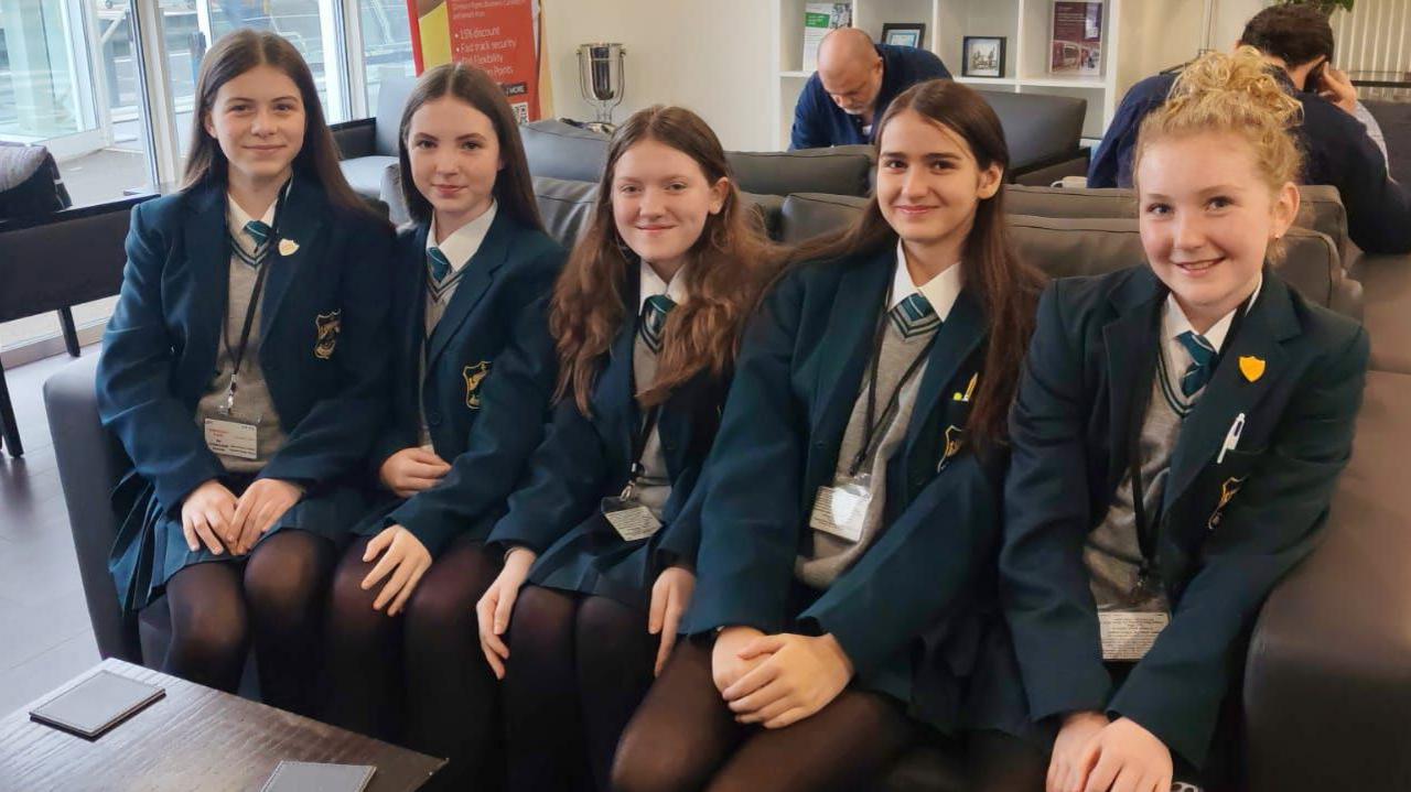 Five girls in their school uniforms sitting on a sofa. They all wear their school uniforms -a teal jacket with grey jumper, teal stripe round the collar and teal tie. 