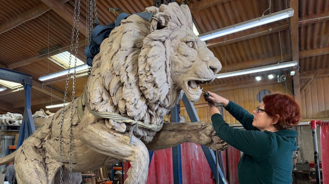 A large lion that is being sculpted out of clay by a woman who is standing in front of it. The woman has short, red hair and is wearing a dark green top. She is holding a tool near the lion's mouth. The statue is in a large studio.