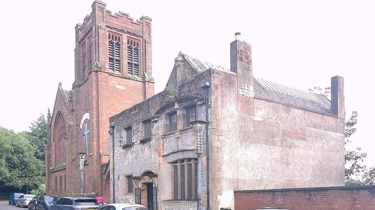 A large re brick church building, with cars parked in front and greenery behind it