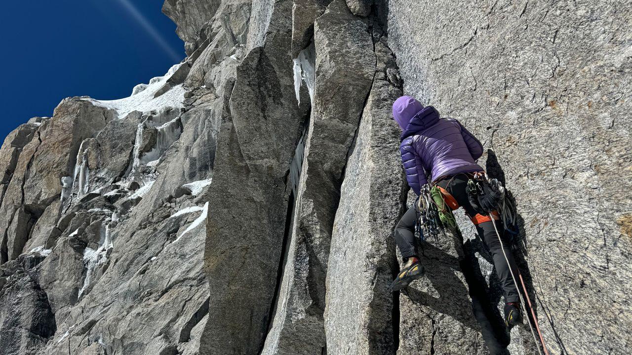 A woman dressed in purple with her back to the camera is climbing the side of a vast grey mountain with blue skies in the distance