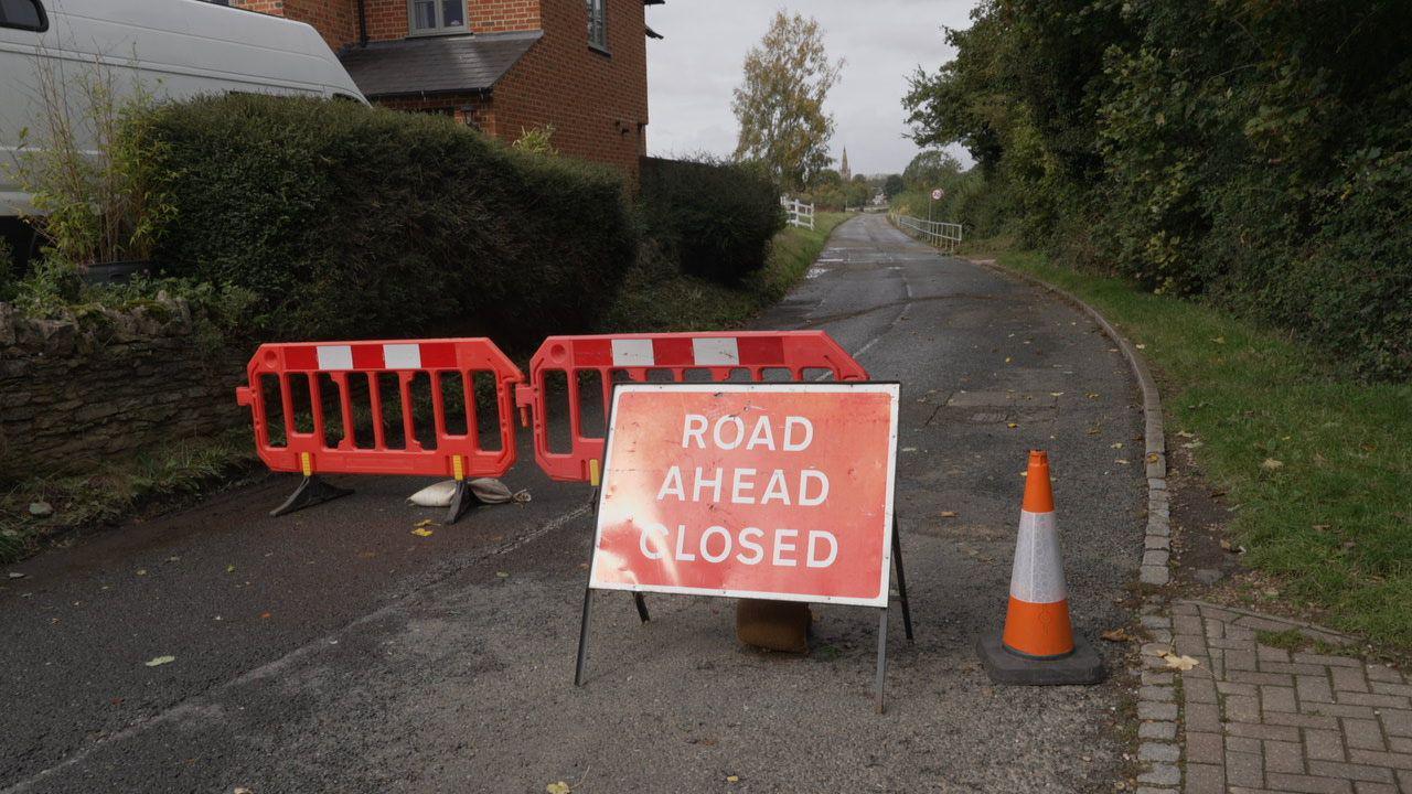 A red road closure sign and a pair of barriers block access to a closed rural road.