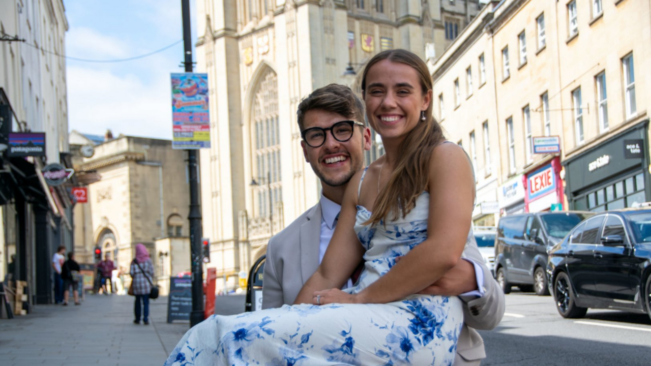 Kirsty sat on Zander's lap, as they both smile on a street in front of a university building