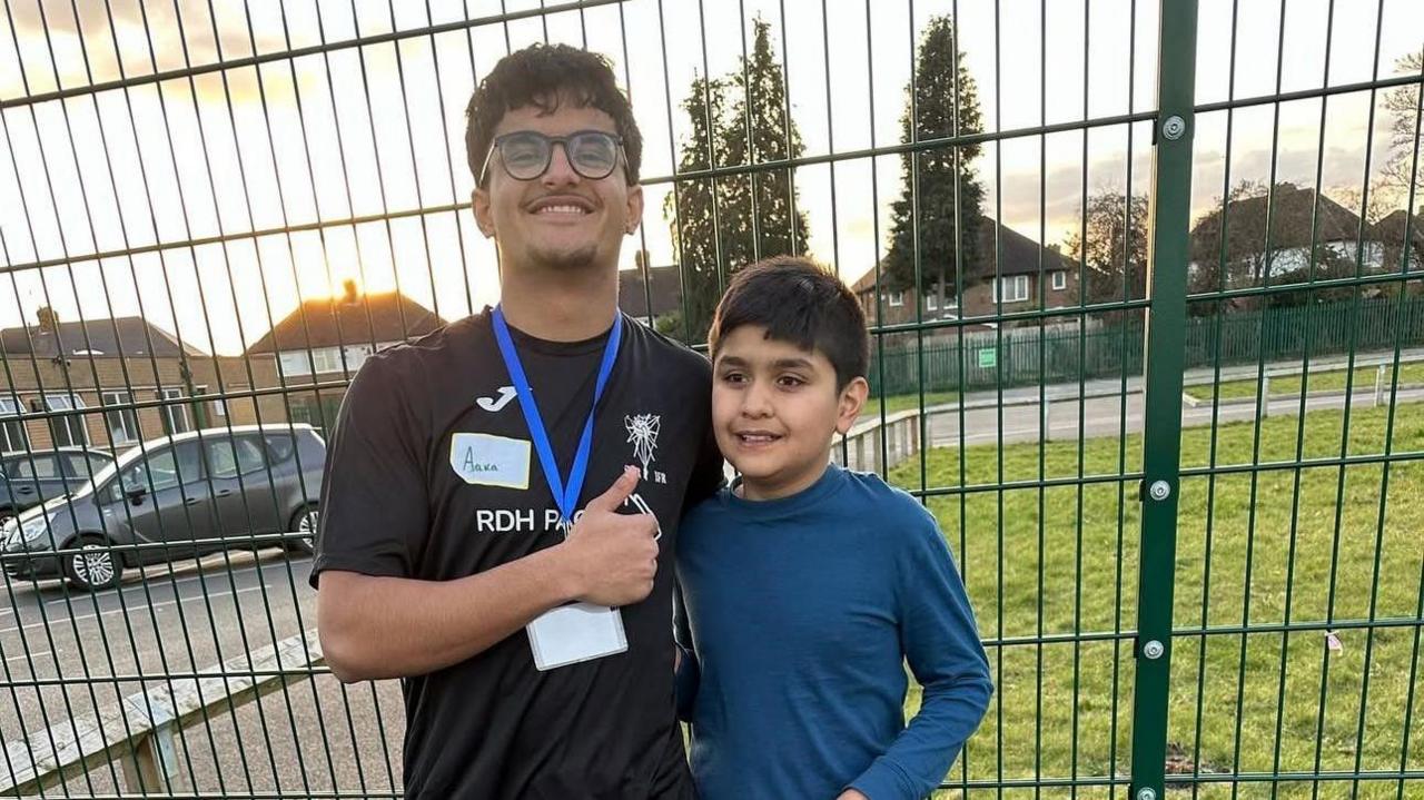 A coach stands with a child and is giving a thumbs up. The coach is wearing a black t-shirt and blue lanyard while the child is wearing a blue long sleeved t-shirt. They are standing in front of a fence.