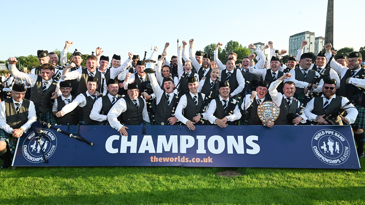 The Inveraray & District Pipe Band celebrating in front of a big World Pipe Band Championships banner saying Champions. They are all in their band uniform and some of them have their hands in the air