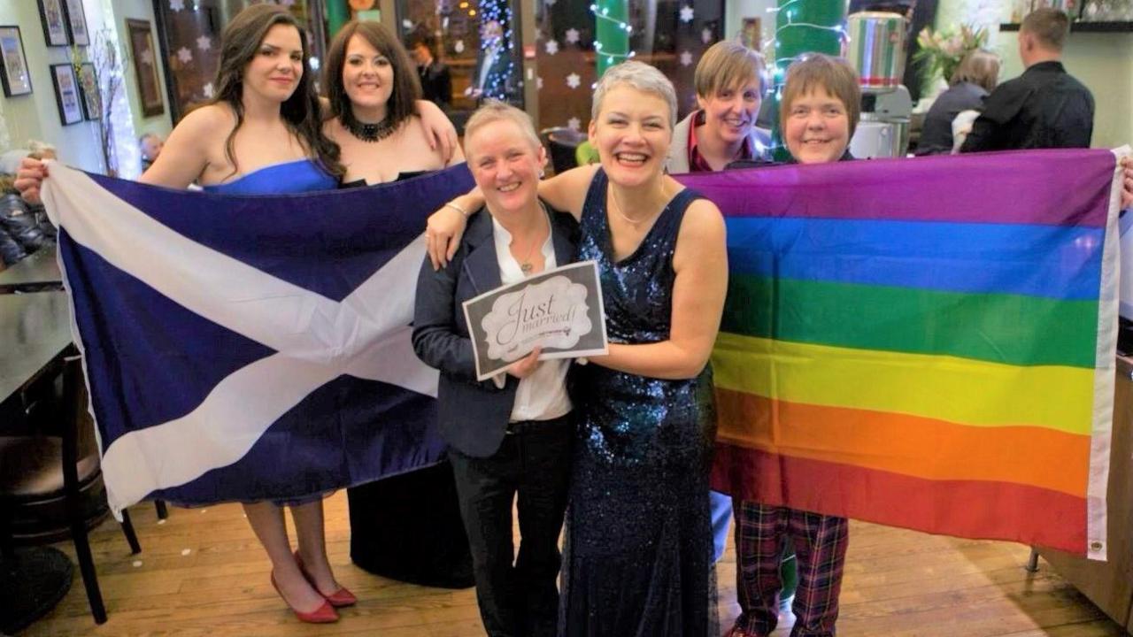 Gerrie and Susan Scott-Douglas smiling in front of a saltire and LGBT+ flag being held up by four women.