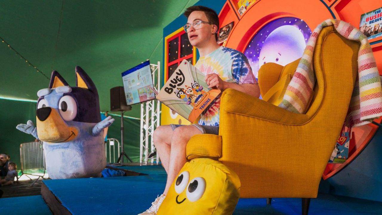 A person sitting in an orange armchair on a stage reading from a book entitled Bluey. On the floor next to them is a giant Bluey the dog stuffed toy.