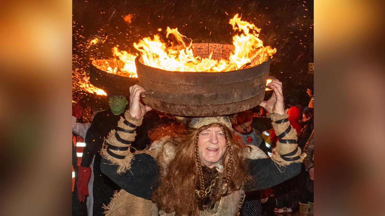 A man, dressed as a viking, leads a procession of people carrying a barrel on their heads. Flames rise out of the barrels.