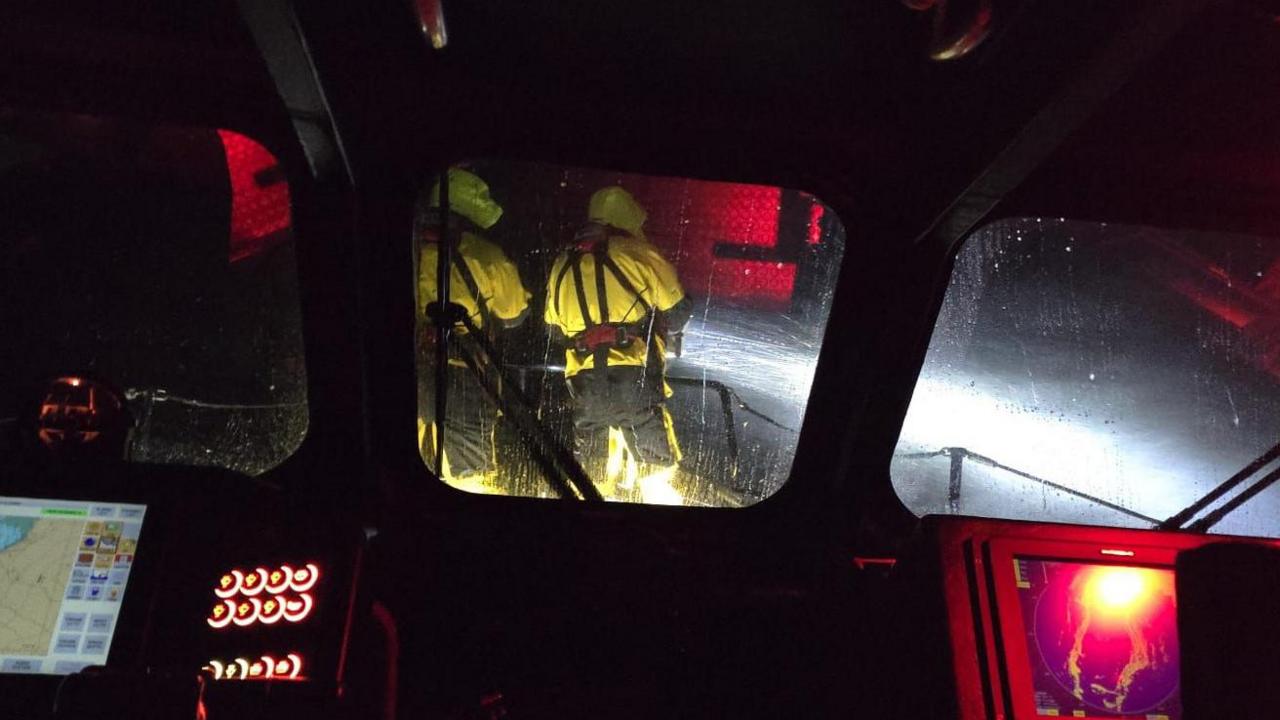 Inside a lightboat in darkness with red illuminated control panels looking out towards two sailors on the bow of the boat