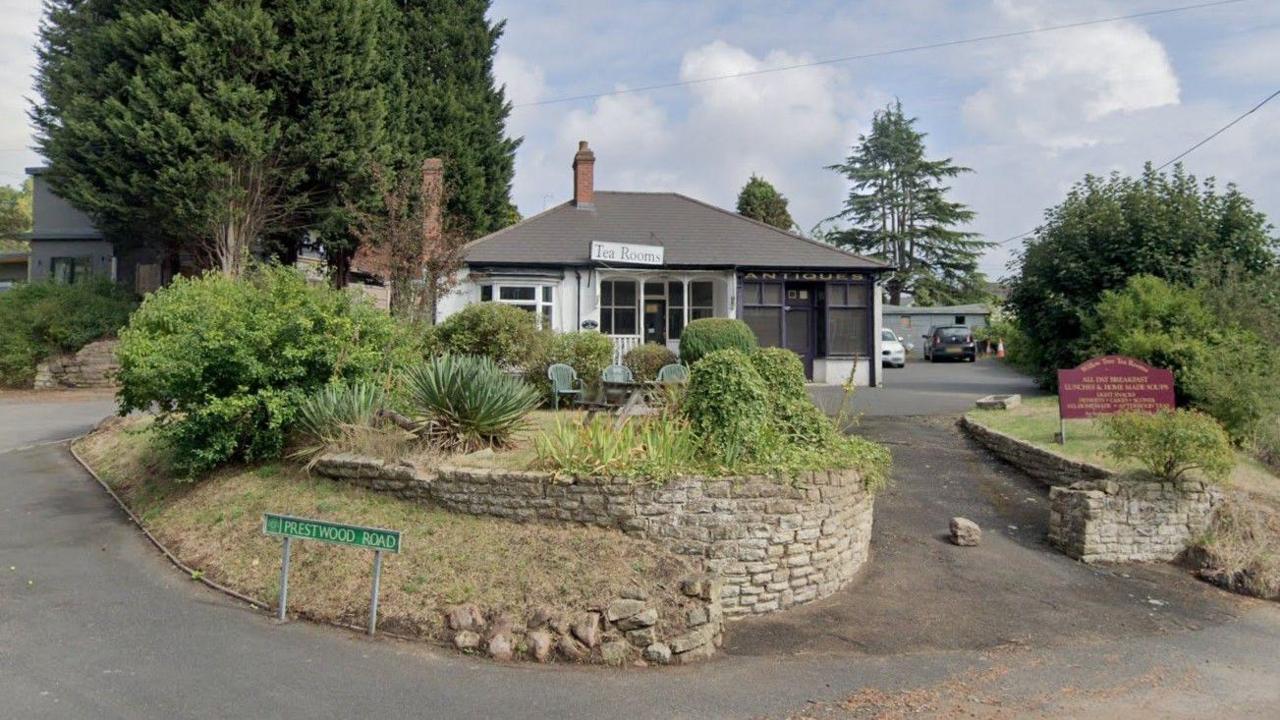 A tea room, a small single-storey building with a veranda at the front, can be seen at the top of a driveway with a brick wall surrounding a raised lawn area which has seating on it.