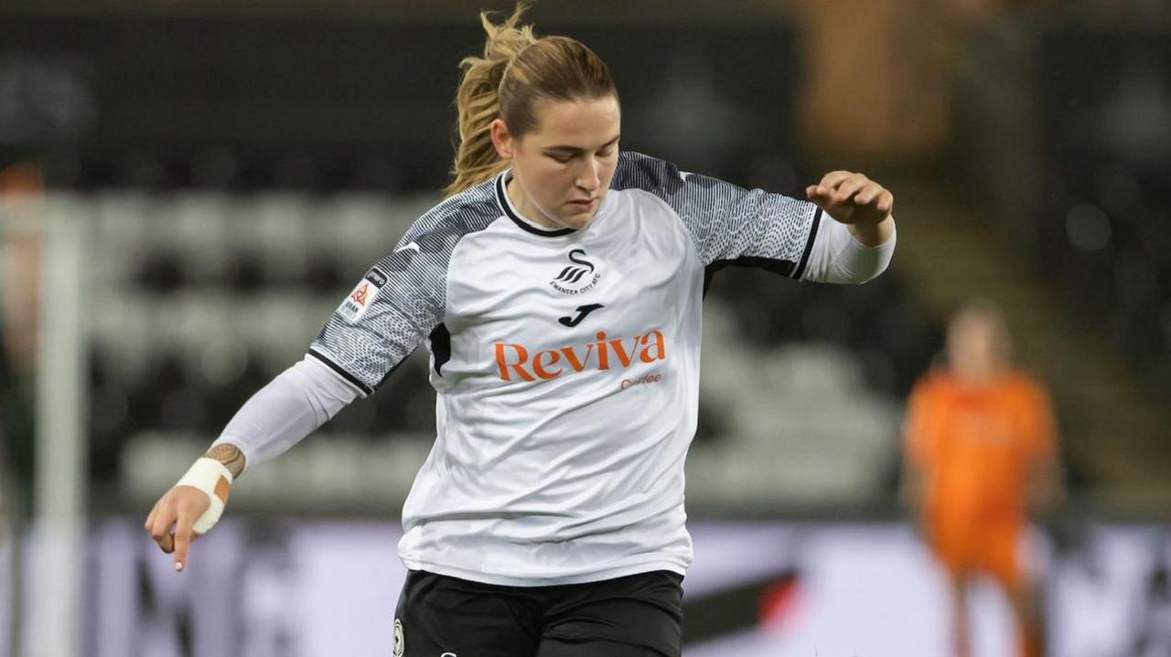 Chloe is wearing black and white Swansea football kit. There is a player in orange blurred in the background. It is a shot of her in action playing football.