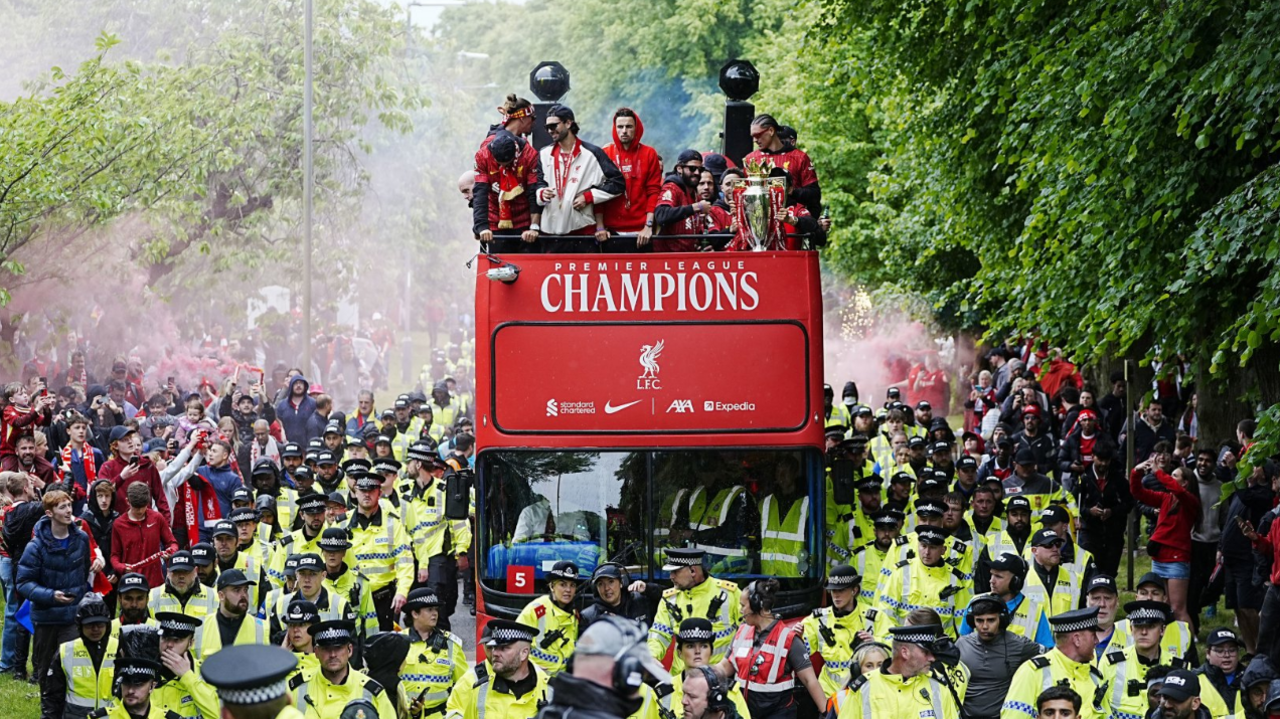 Liverpool's open-top bus moves along flanked by police. Liverpool football players are seen on the top of the open-bus.