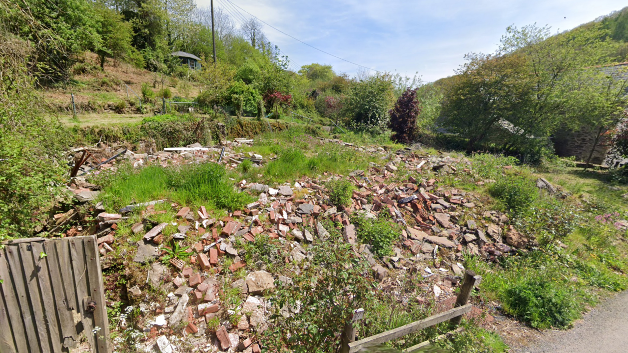 An image taken from Google street view showing an empty and overgrown plot in the countryside. There is a large pile of bricks and building materials lying where the home once stood, with weeds growing between the debris. The plot is surrounded by hills, trees and greenery on a sunny day.