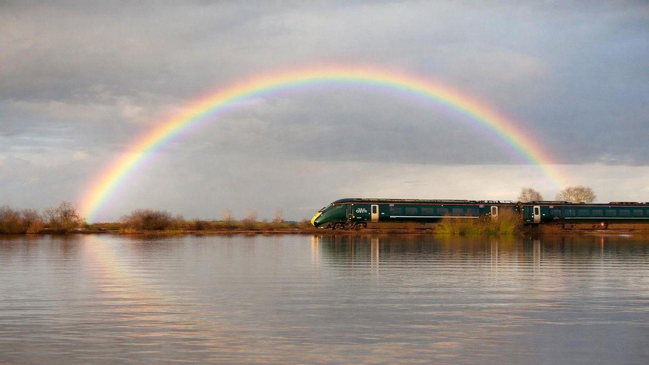 A train passes behind a body of water and in front of a rainbow