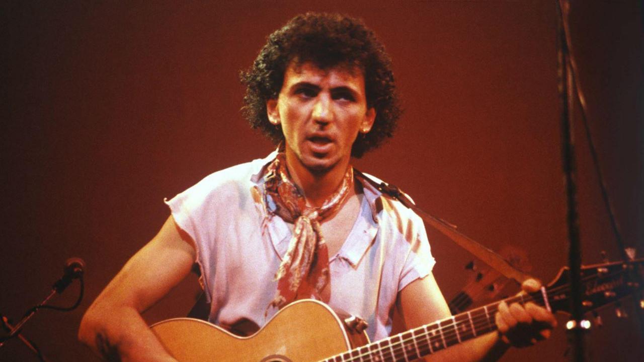 A man with black curly hair and a wooden guitar with a white shirt and red neckerchief