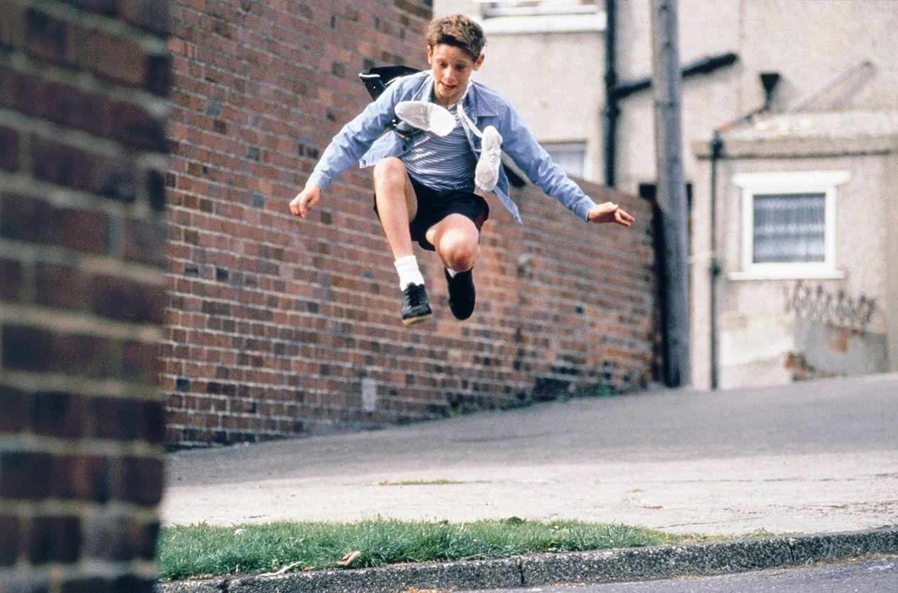 Character Billy Elliot jumping in the air while holding ballet shoes tied around his neck, against an urban backdrop