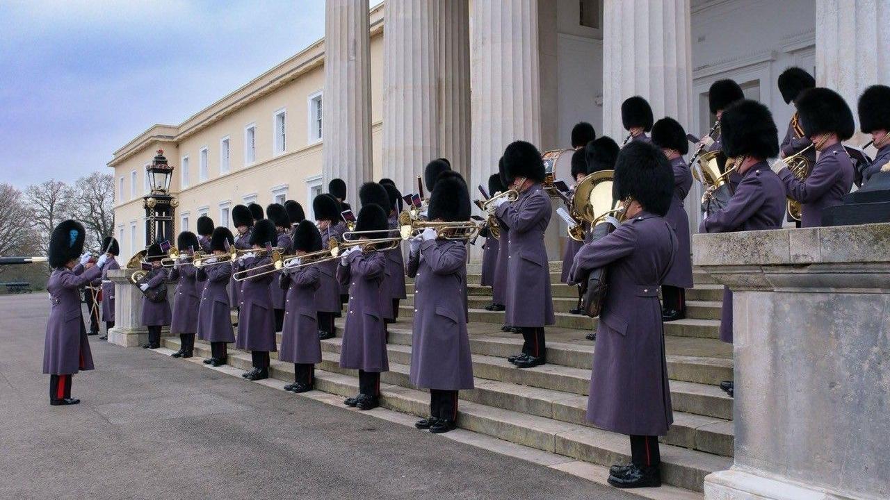 Captain Claire Lawrence, conducting a group of musicians on steps at Sandhurst. They are all wearing bearskin hats and grey coats and trousers, with black shoes. They are all playing musical instruments. 