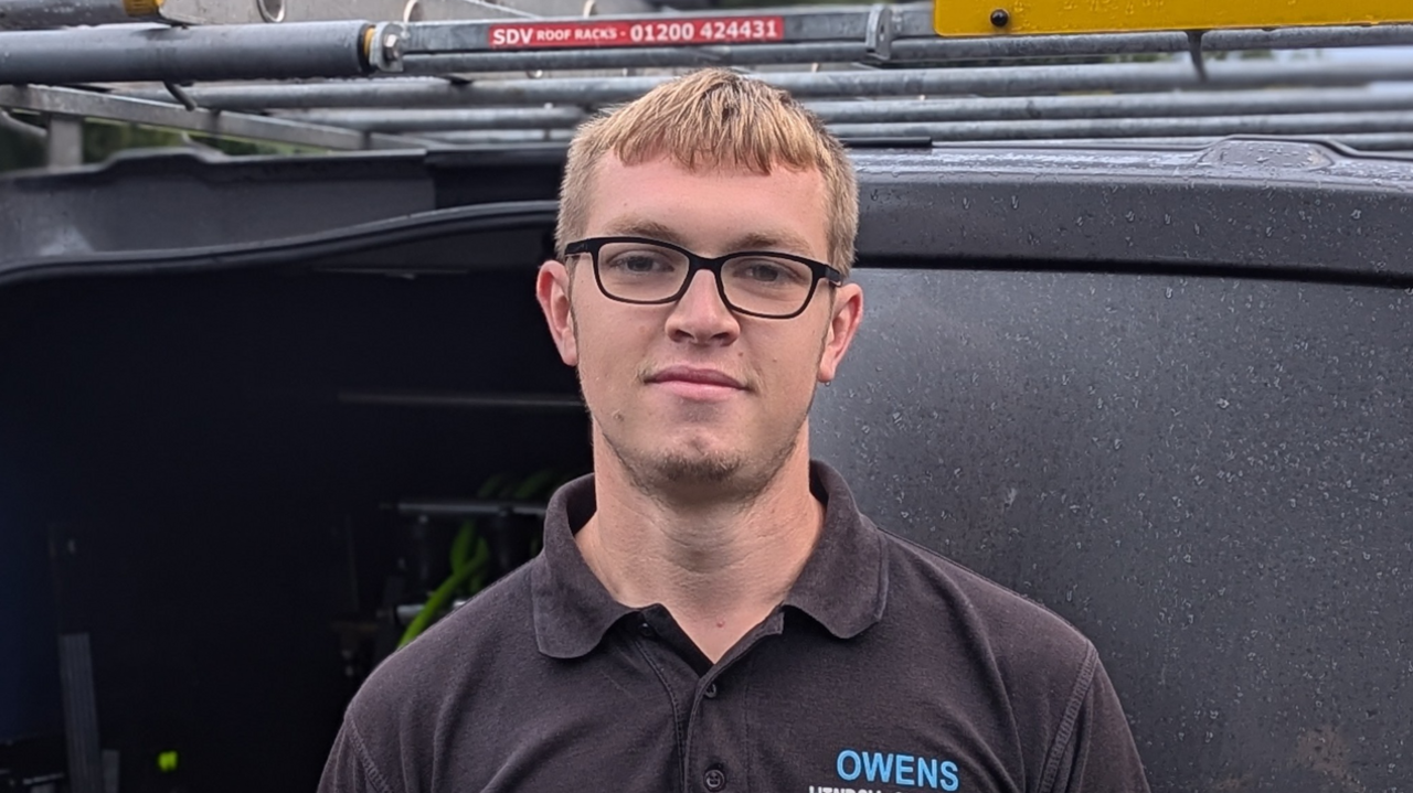 A man with blonde hair and glasses with a black polo shirt stood in front of a black van