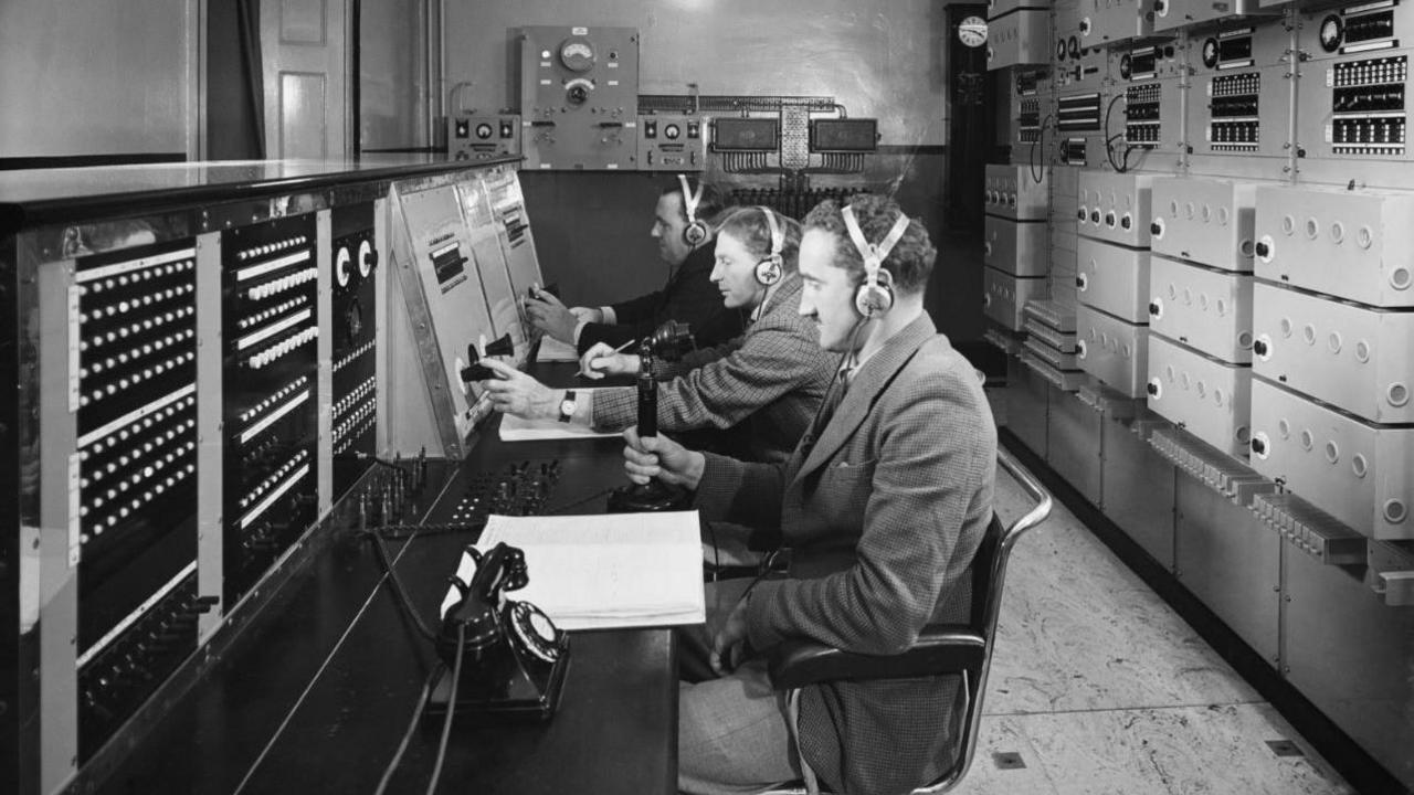 A black and white image of the control room. Three men are sitting at a desk wearing headphones, looking towards a switchboard in front of them with lots of buttons on it. Behind them there is a wall filled with more equipment and wires.