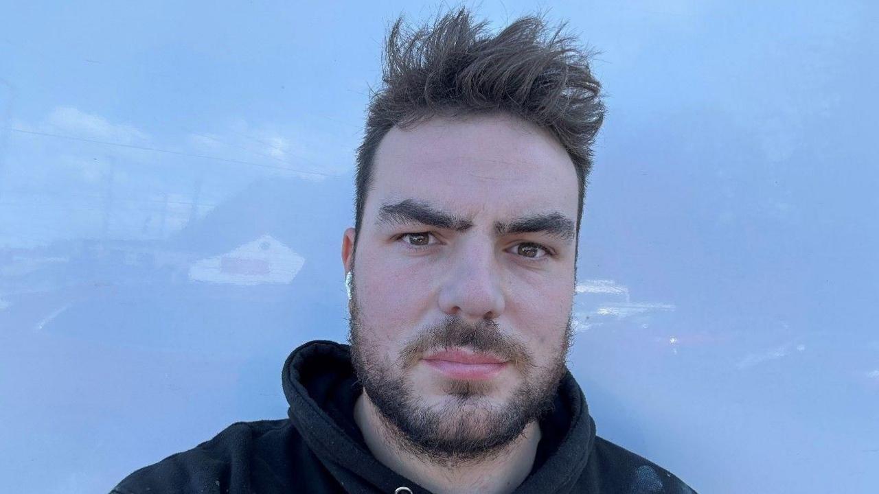 A man with short brown hair and a beard is wearing a black hoodie and is looking towards the camera. There are faint reflections visible on a light blue background.