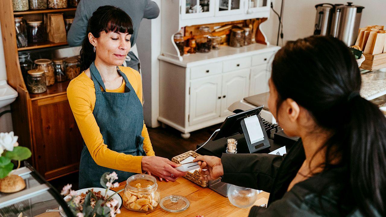 A barista wearing a yellow jumper and a blue overall takes the payment card of a customer wearing a black jacket, in a coffee shop.