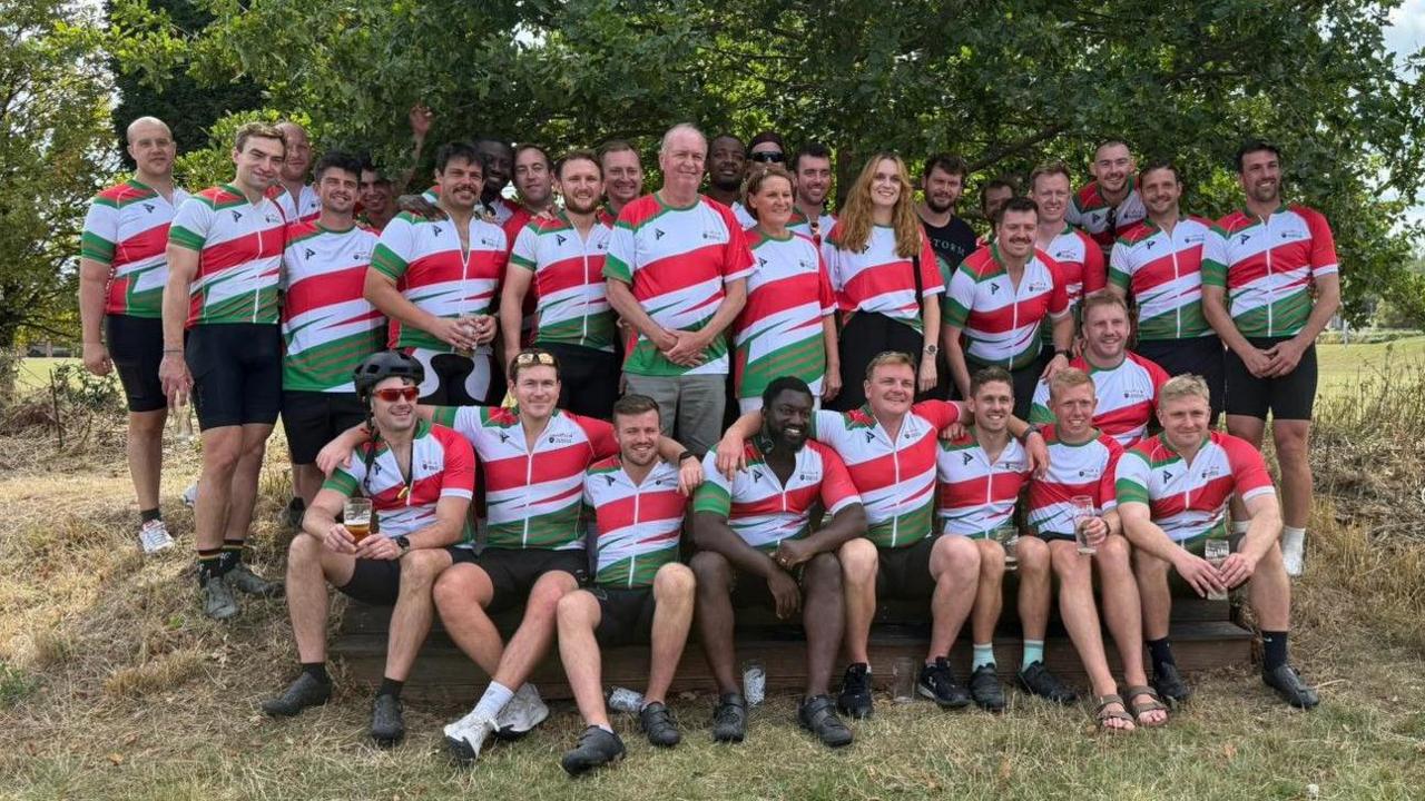 A large group of male and female cyclists, some 40 strong, line up for a group picture at the end of a charity bike ride. They are all wearing the same cycling kit of black shorts and white tops with red and green stripes across them. They are in an open grassy area and gathered under a large tree.